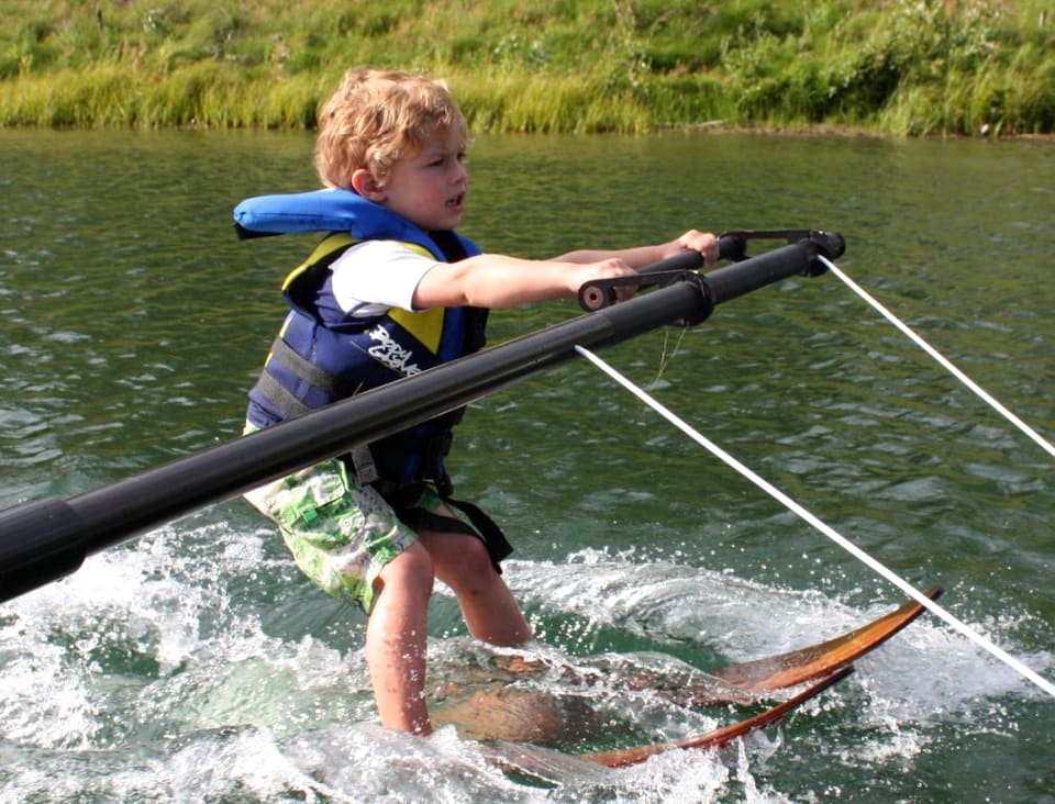 A young skier learning on the boom beside the boat, coach within arm's reach